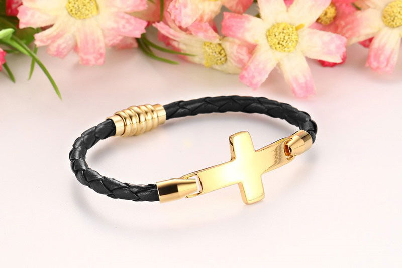 Black braided bracelet with gold cross charm on a light pink background with flowers.