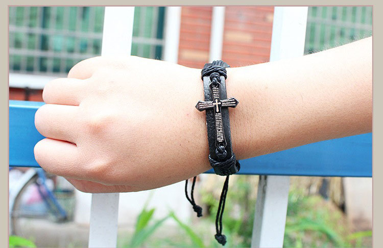 Hand wearing a black leather bracelet with a cross charm against a blurred outdoor background