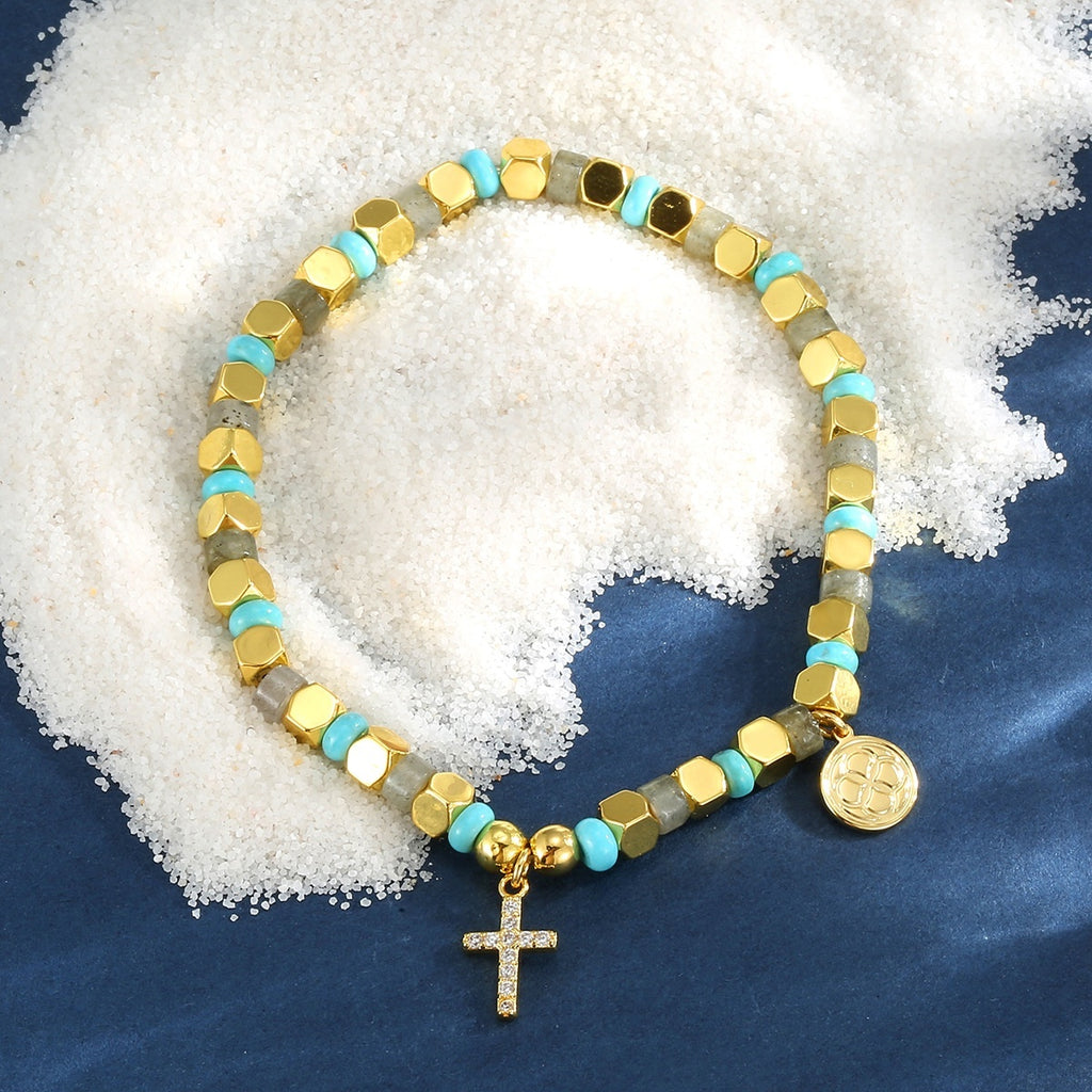 Beaded bracelet with gold accents and a cross charm on a textured white and blue background